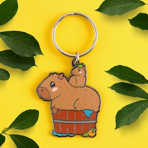 May include: A silver key chain with a cartoon capybara and a smaller capybara riding on its back. The capybaras are in a wooden tub with blue water and a small orange fruit.