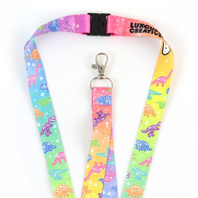 May include: A rainbow lanyard with a dinosaur skeleton pattern and the text "Luxe Creative" printed on it. The lanyard has a silver clip and a black plastic buckle.