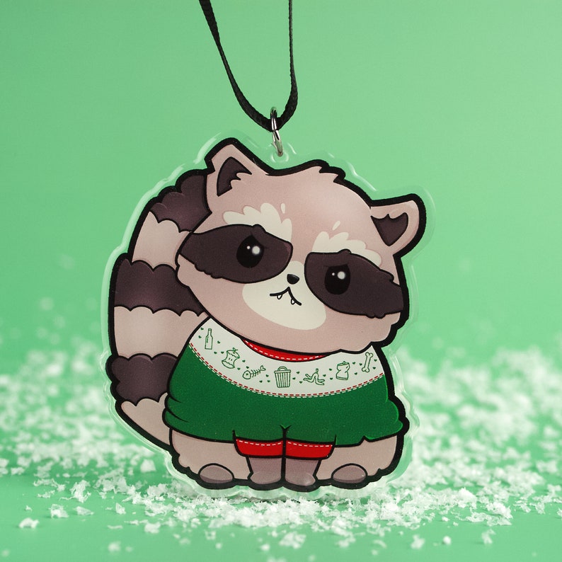 Puede incluir: Un adorno navide&ntilde;o con un mapache de dibujos animados que lleva un su&eacute;ter verde con un dise&ntilde;o festivo. El mapache tiene la nariz negra y los ojos negros. El adorno est&aacute; hecho de pl&aacute;stico transparente y tiene una cuerda negra para colgar.