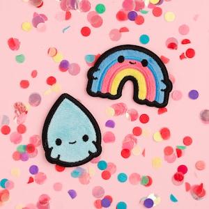 May include: Two embroidered patches on a pink background with colorful confetti. One patch is a smiling rainbow with blue, pink, yellow, and black details. The other is a light blue teardrop shape with a smiling face and black outline.