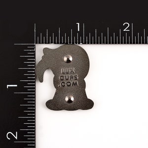 May include: A dark gray enamel pin with a stylized bird design. The pin has two metal clasps on the back and the text "LUX CUPS .COM". The pin is approximately 1 inch in height.