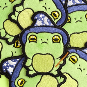 May include: A green frog wearing a blue wizard hat with white stars and a crescent moon. The frog is holding a brown stick in its right hand. The frog is embroidered on a green fabric.