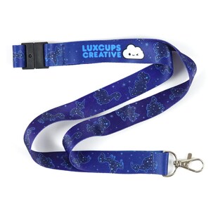 May include: A blue lanyard with a repeating pattern of dinosaur constellations and stars. The lanyard has a black plastic clasp and a silver metal clip. The text "LUXCUPS CREATIVE" is printed on the lanyard, along with a white cloud graphic.