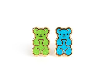 May include: A pair of gold-toned stud earrings, each shaped like a gummy bear. One earring is bright green, the other is blue. Both have a glittery texture and black facial features. The earrings are set against a white background.