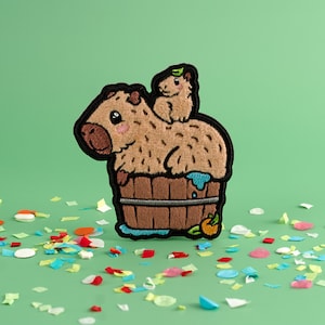 May include: A brown and white embroidered patch of two capybaras, one sitting on top of the other, in a wooden tub. The capybaras are smiling and have small green leaves on their heads. The tub is brown and has a blue rim. The patch is surrounded by colorful confetti.