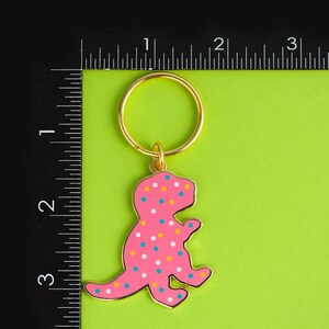 May include: A pink dinosaur keyring with white and yellow polka dots. The keyring has a gold ring and is attached to a green background.