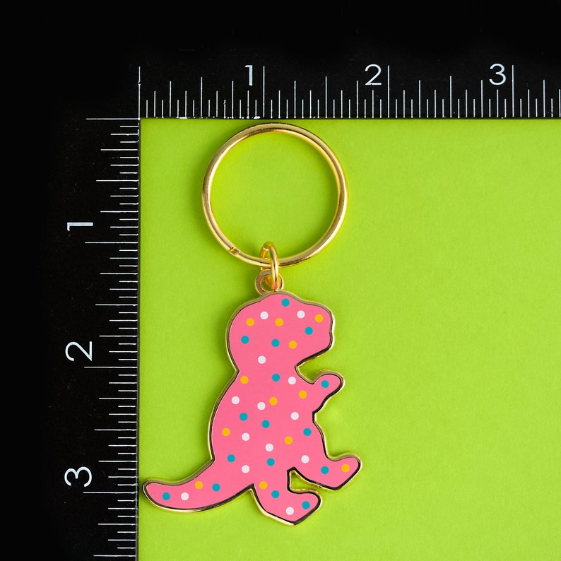 May include: A pink dinosaur keyring with white and yellow polka dots. The keyring has a gold ring and is attached to a green background.
