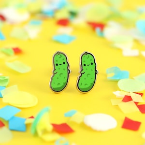 May include: A pair of gold-tone earrings shaped like green pickles with smiling faces. The earrings are on a yellow background with confetti.