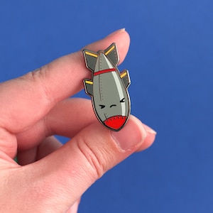 May include: A gray and red enamel pin shaped like a cartoon bomb with yellow wings. The bomb has a frowning face.