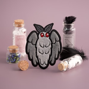 May include: A gray felt embroidered patch of a moth with red eyes. The moth has large wings and a fuzzy body. The patch is surrounded by small glass bottles with labels that say "Eyes" and "Ravens".