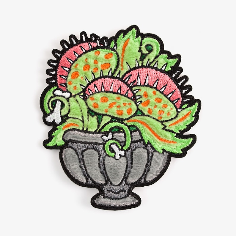 May include: A green and pink embroidered patch of a Venus flytrap plant in a gray pot. The plant has orange spots and is in a gray pot with a white bone design.