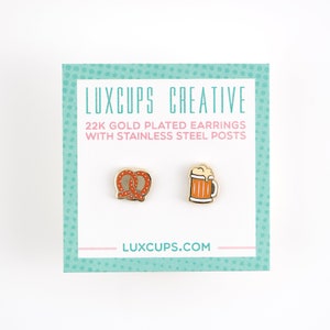 May include: A pair of gold-plated earrings featuring a pretzel and a beer mug. The earrings are on a white background with a pink and teal border. The text "LUXCUPS CREATIVE" and "22K GOLD PLATED EARRINGS WITH STAINLESS STEEL POSTS" is printed on the card. The text "LUXCUPS.COM" is printed on the bottom of the card.