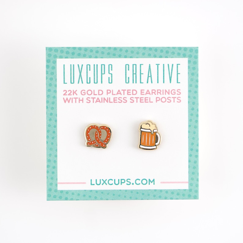 May include: A pair of gold-plated earrings featuring a pretzel and a beer mug. The earrings are on a white background with a pink and teal border. The text "LUXCUPS CREATIVE" and "22K GOLD PLATED EARRINGS WITH STAINLESS STEEL POSTS" is printed on the card. The text "LUXCUPS.COM" is printed on the bottom of the card.