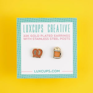 May include: A pair of gold-plated earrings featuring a pretzel and a beer mug. The earrings are on a white card with the text "LUXCUPS CREATIVE 22K GOLD PLATED EARRINGS WITH STAINLESS STEEL POSTS LUXCUPS.COM".