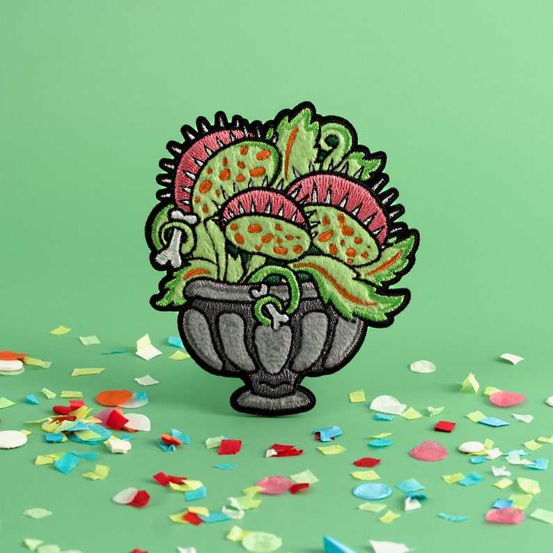 May include: A green, pink, and orange embroidered patch of a carnivorous plant in a gray pot. The patch is on a green background with confetti.
