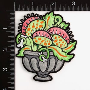 May include: A green, pink, and orange embroidered patch of a carnivorous plant in a gray pot. The plant has multiple mouths with teeth and orange spots.