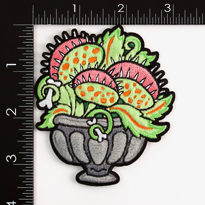 May include: A green, pink, and orange embroidered patch of a carnivorous plant in a gray pot. The plant has multiple mouths with teeth and orange spots.