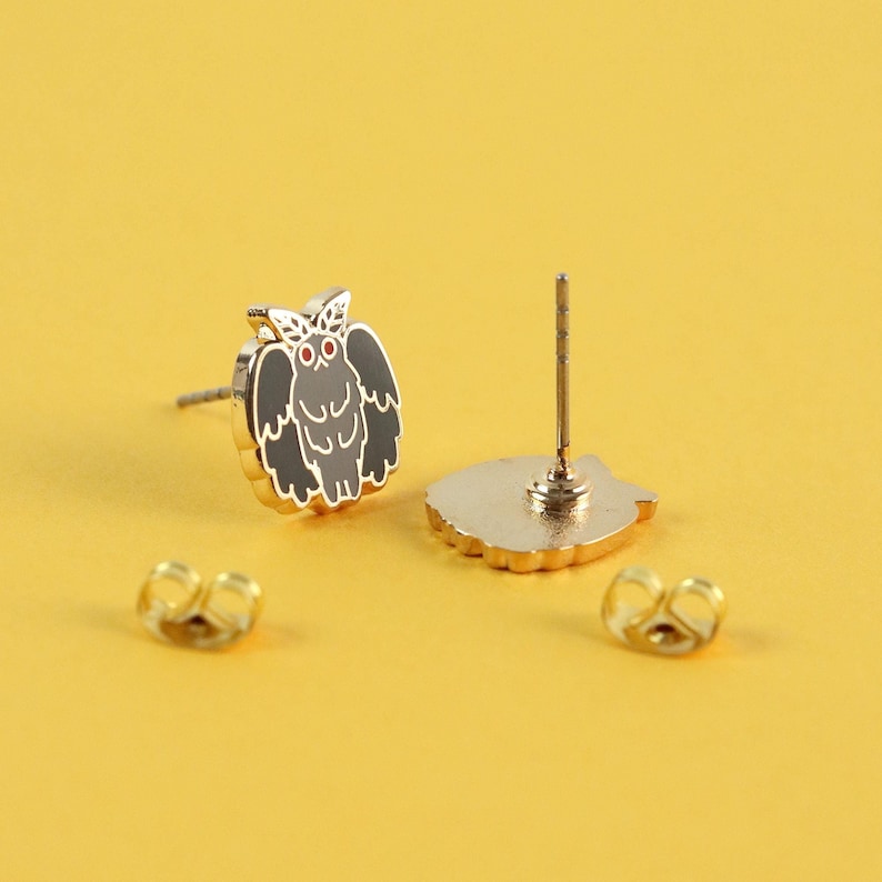 K&ouml;nnte beinhalten: Goldfarbene Eulen-Ohrstecker mit schwarzen und grauen Emaille-Details und roten Augen. Das Eulen-Design hat eine goldene Umrandung und ist vor einem gelben Hintergrund platziert. Enth&auml;lt zwei goldene Ohrringverschl&uuml;sse.
