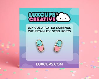 May include: A pair of 22k gold plated earrings with stainless steel posts shaped like small capsules with a pink bottom and blue top, each featuring a smiling face. The earrings are displayed on a card labeled LUXCUPS CREATIVE.