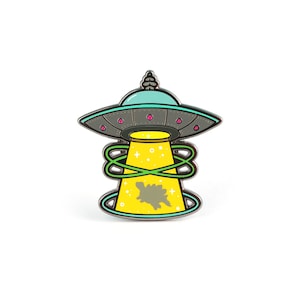 May include: A silver enamel pin featuring a cartoon UFO with a green beam of light shining down on a gray dinosaur. The UFO is blue and gray with pink accents. The background is white.