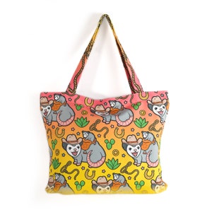 May include: A yellow and pink tote bag with a repeating pattern of cartoon opossums wearing cowboy hats and riding other opossums. The opossums are surrounded by stars, horseshoes, and cacti.