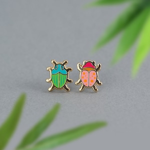 May include: A pair of gold-tone earrings featuring a green and blue beetle and a pink and orange beetle.