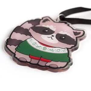 Puede incluir: Un mapache de dibujos animados rosa y gris que lleva un su&eacute;ter navide&ntilde;o verde con ribete blanco y fondo rojo. El su&eacute;ter tiene un patr&oacute;n de basura, huesos y otros objetos. El mapache tiene la nariz negra y los ojos negros. El mapache sostiene una cinta negra.