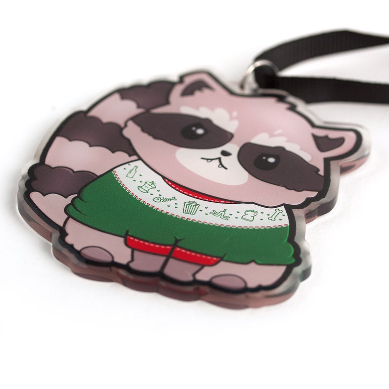 Puede incluir: Un mapache de dibujos animados rosa y gris que lleva un su&eacute;ter navide&ntilde;o verde con ribete blanco y fondo rojo. El su&eacute;ter tiene un patr&oacute;n de basura, huesos y otros objetos. El mapache tiene la nariz negra y los ojos negros. El mapache sostiene una cinta negra.