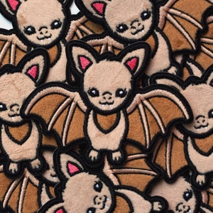 May include: A collection of brown plush bat patches with black embroidered details. The bats have large eyes, small fangs, and outstretched wings.