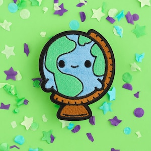 May include: A green and blue embroidered globe with a smiling face, surrounded by confetti on a green background.