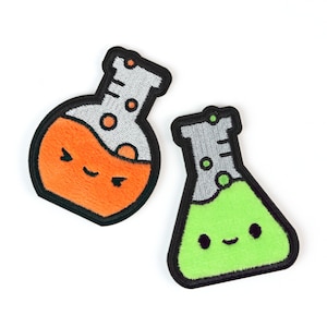 May include: Two embroidered patches shaped like laboratory flasks. One round-bottom flask contains orange liquid with a smiling face, and one conical flask contains green liquid with a smiling face. Both have black borders and gray necks.