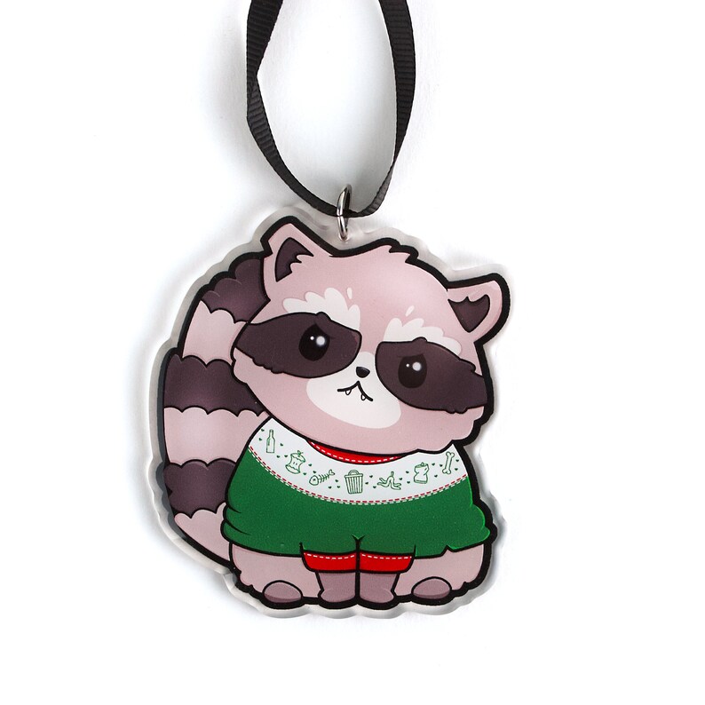 Puede incluir: Un mapache rosa y gris con un su&eacute;ter navide&ntilde;o verde con ribete rojo y un patr&oacute;n blanco de iconos navide&ntilde;os. El adorno est&aacute; hecho de acr&iacute;lico transparente y tiene una cinta negra.