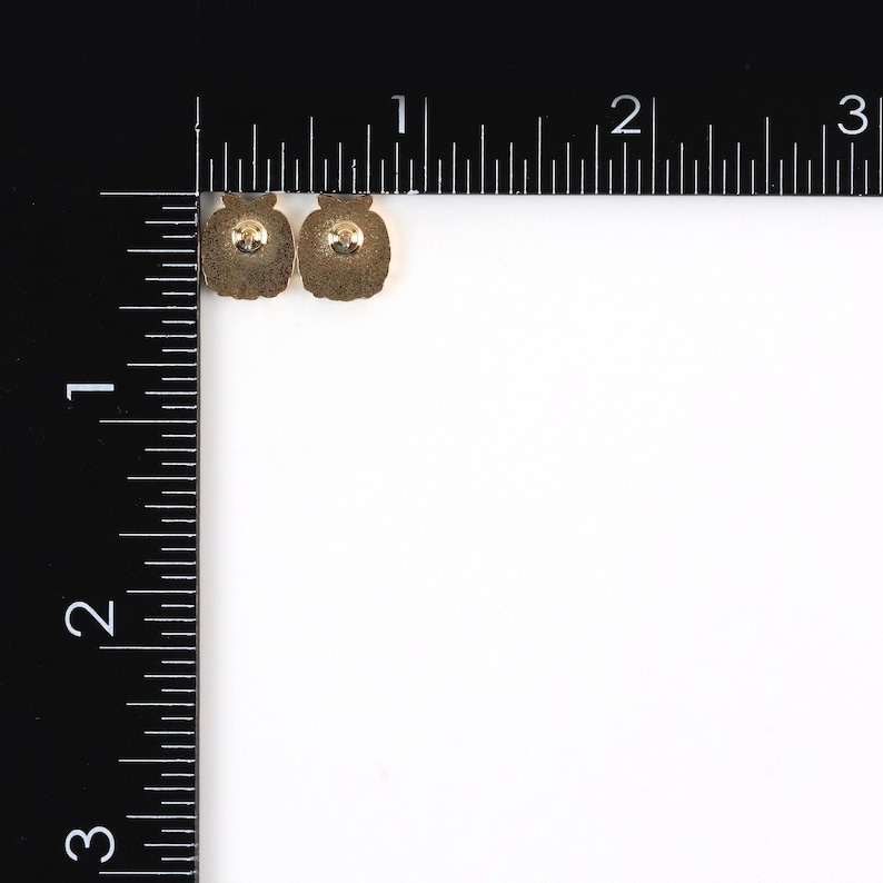 K&ouml;nnte beinhalten: Zwei kleine, goldfarbene Ohrringe mit einer abgerundeten, gl&auml;serartigen Form. Jeder Ohrring hat einen Stift auf der R&uuml;ckseite zur Befestigung am Ohr. Die Ohrringe sind vor einem wei&szlig;en Hintergrund mit einem schwarzen Lineal platziert.