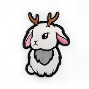 May include: Embroidered patch featuring a white bunny with brown antlers, a gray chest, and pink inner ears. The patch has a black border and a slightly grumpy expression. This whimsical design is perfect for adding to clothing or accessories.