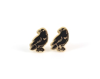 May include: A pair of gold-tone earrings with black raven silhouettes. The ravens are facing to the right and have a detailed design.