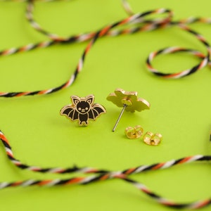 May include: A pair of gold-tone stud earrings featuring a black bat design. The earrings are on a green surface with black and white string.
