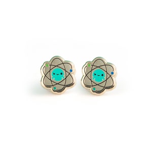 May include: A pair of silver-toned earrings with a cartoon atom design. The atom has a turquoise center with a smiling face and green and blue orbiting electrons.