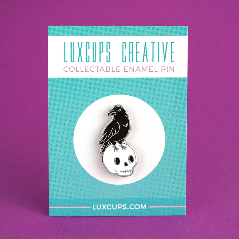 Może przedstawiać: Czarna emaliowana szpilka z krukiem siedzącym na białej czaszce. Szpilka znajduje się na białej kartce z tekstem "LUXCUPS CREATIVE COLLECTABLE ENAMEL PIN" i "LUXCUPS.COM".