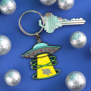 May include: A silver key and a keychain with a cartoon UFO design. The UFO is gray, teal, and yellow, with a green beam and a dinosaur silhouette. The keychain is attached to a silver key ring. The background is blue with silver glitter balls.