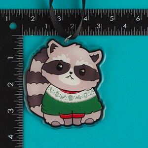 Puede incluir: Un adorno navide&ntilde;o con un mapache de dibujos animados que lleva un su&eacute;ter verde con ribete rojo y un patr&oacute;n blanco de botes de basura, huesos y botellas de cerveza. El mapache tiene la nariz rosa y los ojos negros.