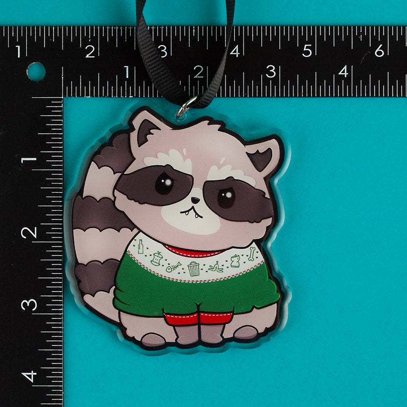 Puede incluir: Un adorno navide&ntilde;o con un mapache de dibujos animados que lleva un su&eacute;ter verde con ribete rojo y un patr&oacute;n blanco de botes de basura, huesos y botellas de cerveza. El mapache tiene la nariz rosa y los ojos negros.