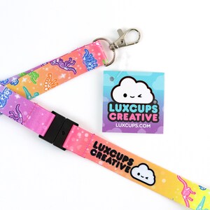 May include: A pink, yellow, and blue lanyard with a rainbow gradient and a white cloud with a winking face. The lanyard has a black plastic clip and a silver metal swivel hook. The lanyard has a repeating pattern of white dinosaur skeletons on a colorful background. The lanyard also has the text "LUXCUPS CREATIVE" and the website "LUXCUPS.COM" printed on it.