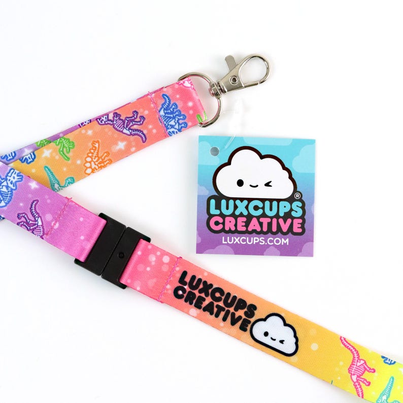 May include: A pink, yellow, and blue lanyard with a rainbow gradient and a white cloud with a winking face. The lanyard has a black plastic clip and a silver metal swivel hook. The lanyard has a repeating pattern of white dinosaur skeletons on a colorful background. The lanyard also has the text "LUXCUPS CREATIVE" and the website "LUXCUPS.COM" printed on it.