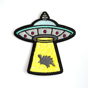 May include: Embroidered patch of a grey and black UFO with a yellow light shining down on a grey dinosaur. The UFO has a teal rim and red lights.