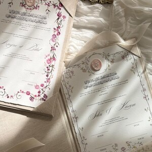 Op de afbeelding: Twee off-white huwelijkscontractdocumenten met bloemenranden, roze waszegels en gedrukte tekst inclusief de namen Maryne et Bilal en Selin & Kareem. Een houten waszegelstempel rust op een klein versierd gouden dienblad.