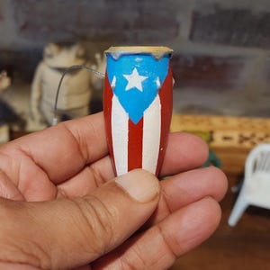 May include: A miniature drum, painted with the flag of Puerto Rico. The drum features a blue section with a white star, red stripes, and white stripes. The drum is held in a hand.