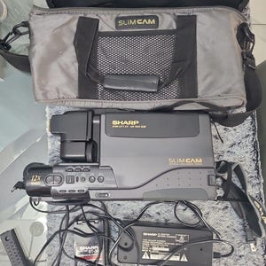 May include: A grey Sharp Slimcam video camera with a 12x zoom lens, accompanied by a matching grey carrying bag, a Sharp BT-30 battery pack, and a black Sharp AC adapter with power cords. The camera features the text Slimcam and Sharp.