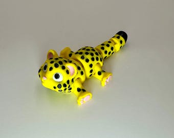 May include: A yellow toy leopard with black spots, pink ears, and pink paws. The leopard has a black-tipped tail and is lying down. The toy is made of plastic and is likely a child's toy.
