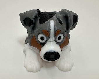 May include: A 3D-printed dog figurine in a sitting position. The dog has a gray, black, brown, and white color scheme. The dog's eyes are large and round, and its paws are white. The figurine is a decorative item.
