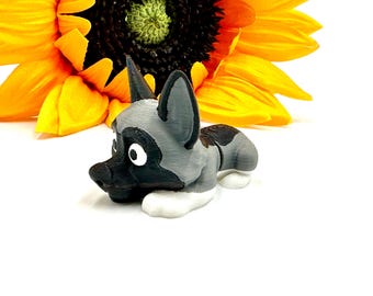 May include: A 3D-printed figurine of a cartoon dog in a lying down position. The dog has a black nose, ears, and markings, with gray and white accents. A bright yellow sunflower is in the background.